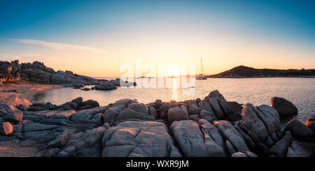Panoramic view of two yachts moored in a bay as the sun sets over the Mediterranean sea and large granite boulders on the coast of Cavallo Island in t Stock Photo