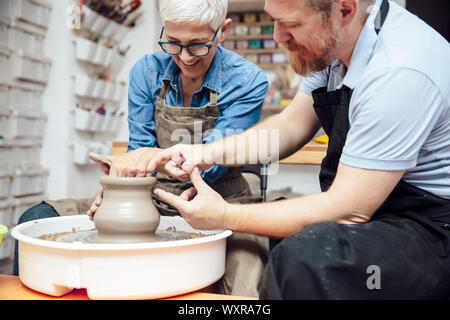 Senior woman spinning clay on a wheel with a help of a teacher at pottery class Stock Photo