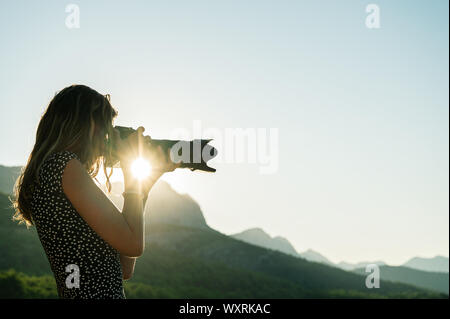 Young female photographer taking photos of beautiful nature using zoom lens on a camera with sunburst of rising sun shining in between her hands. Stock Photo