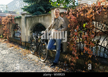 The man is standing by the fence with wild grapes. He is holding the bike. Stock Photo