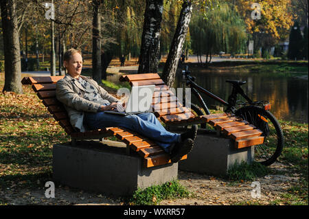The man on a lounger near the lake is working on a laptop. A bike is nearby. Stock Photo