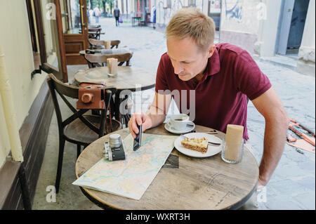 The man is holding a smartphone. He is sitting at the cafe table on the street. On the table there is a vintage camera, a tourist map, a coffee and a Stock Photo
