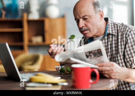 Senior hispanic man eating fresh salad in the morning sitting in the living room near laptop Stock Photo