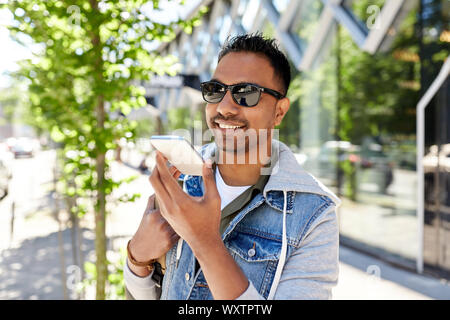 travel, tourism and lifestyle concept - smiling indian man with backpack using voice command recorder on smartphone on city street Stock Photo