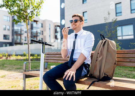 business and people and concept - young smiling businessman with backpack and scooter sitting on street bench and using voice command recorder on smar Stock Photo