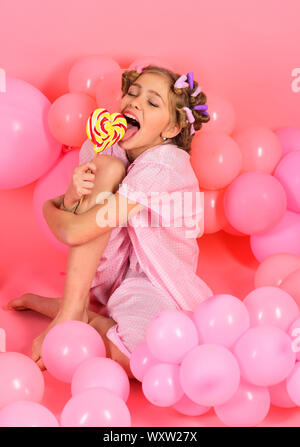 Party balloons, kid in curlers, pajama fashion. Childhood, happiness, sweet dreams. Small girl child eat lollipop on pink. Diet, birthday, punchy past Stock Photo