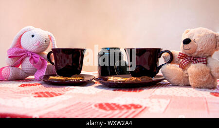 Teddy bears tea party with two cute little plush toys sitting on either side of cups of tea with cookies on a red tablecloth Stock Photo