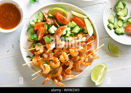Prawns skewers and vegetables salad from sliced cucumber and tomatoes on white plate over wooden table. Grilled shrimp skewers with sauce and fresh ve Stock Photo