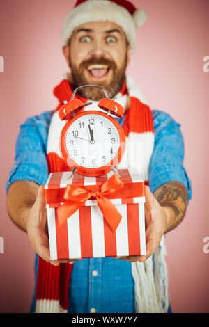 Christmas alarm clock and gift box in blurred man hands. Hipster in santa hat, scarf smile on pink background. Boxing day concept. New year, xmas holi Stock Photo