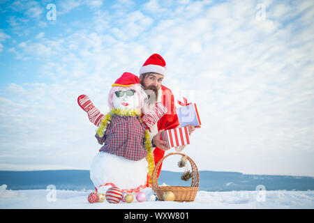 Surprised man opening xmas present box. Snowman in pink wig, mittens and glasses. Hipster in santa hat and red costume on cloudy sky. Christmas and ne Stock Photo