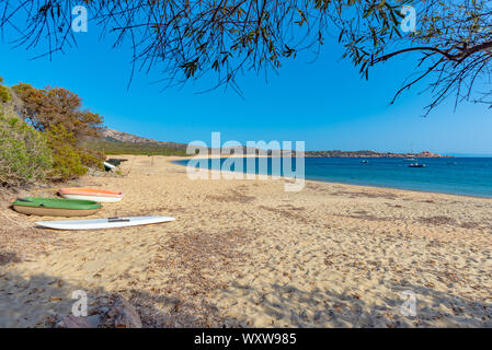 Framed view on a secluded beach with golden sand, blue sea water and rocky, vegetated and granitic hills in the background and few boats in the foregr Stock Photo