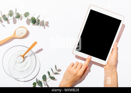 Woman's hands using blank-screen tablet device next to homemade organic cosmetics clay mask on white table among eucalyptus twigs, top view. Stock Photo