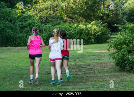 Three girls are training together running in a park on green grass with bushes and trees all around them. Stock Photo