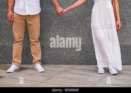 Man in white shirt and woman in white dress holding hands of each other while standing near the grey wall. Stock Photo
