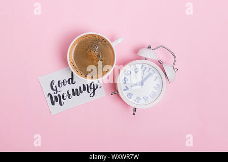 Analog alarm clock, cup of coffee and note with text good morning on pink background. Stock Photo