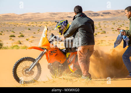 Merzouga, Morocco - February 25, 2016: A motorbike rider churns up sand as he tries to get his orange bike going in the Sahara Desert. Two men are com Stock Photo