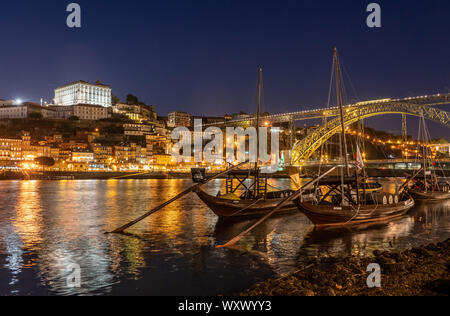 Rabelo boats used to transport port in Porto in Portugal with a panorama of the skyline of the old town at night with lights reflecting in the river Stock Photo