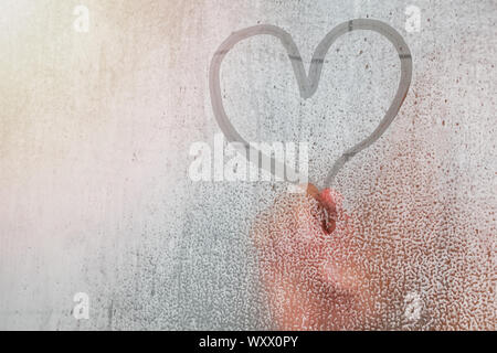 Adult person finger drawing a heart shape on the misted glass with drops in the bathroom. Stock Photo