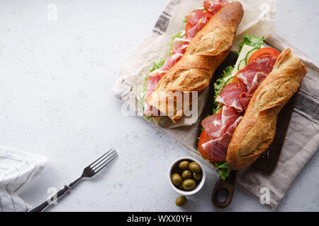 Top view of two fresh baguette sandwiches bahn-mi styled with olives. Ham, sliced cheese, tomatoes and fresh lettuce on dark wooden cutting board on c Stock Photo