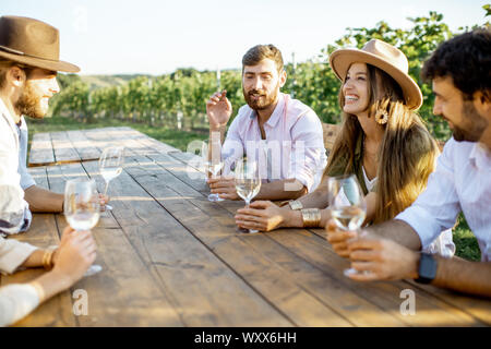 Group of a young people drinking wine and talking together while sitting at the dining table outdoors on the vineyard on a sunny evening Stock Photo