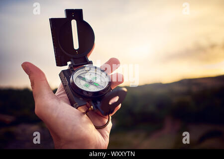 Close-up of a human hand holding an analogue compass in backlight at sunset over hills Stock Photo