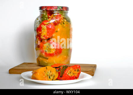 Stuffed with celery, parsnips, carrots and other vegetables, pickled red and yellow peppers lie on a white plate close-up against a closed jar of pepp Stock Photo
