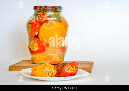 Stuffed with celery, parsnips, carrots and other vegetables, pickled red and yellow peppers lie on a white plate close-up against a closed jar of pepp Stock Photo