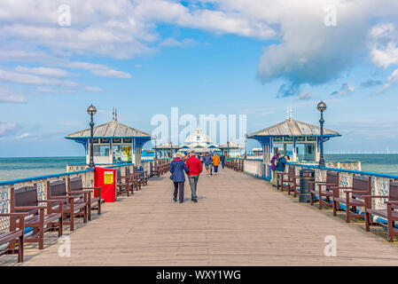 The pier at Llandudno with tourists strolling.  Wind turbines are on the horizon and there is a blue, cloudy sky overhead. Stock Photo