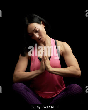 young beautiful athletic girl in a pink top sits with crossed legs in a lotus position, hands makes a gesture of namaste with joined hands in front of Stock Photo