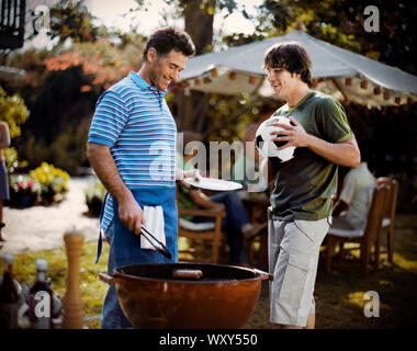 Smiling mature man cooking on a barbeque grill while listening to his teenage son in the back yard of their home. Stock Photo