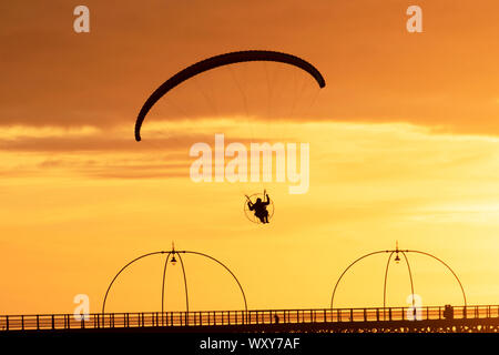 Powered hand glider silhouetted in the evening sun, takes a motorized powered flight in light winds as the sun sets in Southport. Paramotor flying machines & stunt flying as the sun sets; a hand glider silhouette against orange clouds, enjoying flying propeller powered aircraft over the Irish Sea coast. Stock Photo