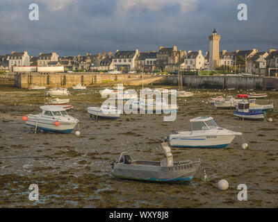 Many small boats sitting in mud at low tide in the harbor at Roscoff, France, with the town and lighthouse lit by the evening sun. Stock Photo