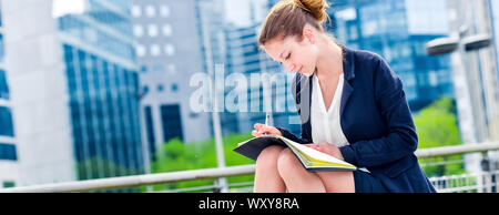 panoramic view on dynamic young executive girl taking notes on her agenda, outside. Symbolizing a job search or a trade of outsourcing Stock Photo