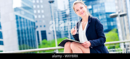 panoramic view on dynamic young executive girl taking notes on her agenda, outside. Symbolizing a job search or a trade of outsourcing Stock Photo