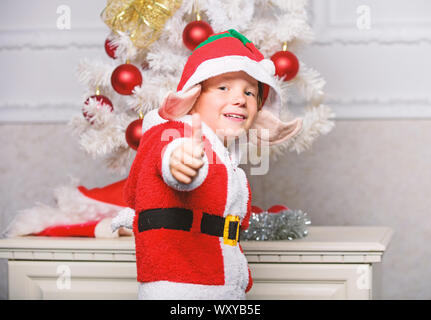 Christmas tree ideas for kids. Boy kid dressed as cute elf magical creature white artificial ears and red hat near christmas tree. Christmas elf costume for child. Christmas party with elf costume. Stock Photo