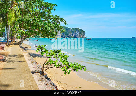 Walk the shady seaside promenade of Ao Nang and watch the golden beach, turquoise waters of Andaman sea, lush tropical greenery and rocky cliffs on ba Stock Photo