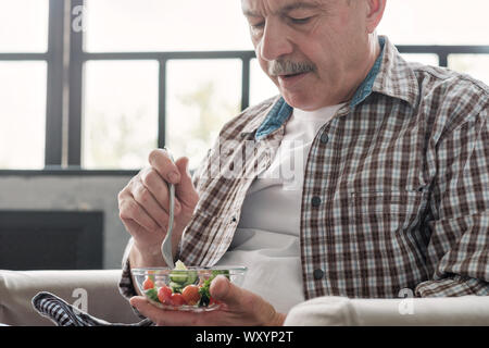 Senior hispanic man sitting in the living room eating fresh vegetable salad Stock Photo