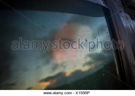 A man sitting in a car looking out. Reflections of the sunset sky on the window.