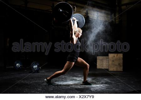 Side view of young woman weightlifting barbell in dark gym