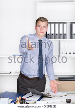 Junger wütender Geschäftsmann steht vor einem Regal im Büro und streckt einem Arm aus in Richtung Kamera. Der Mann schaut zur Kamera.