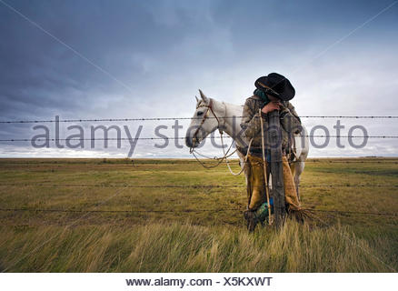 A cowboy standing leaning on a fence post on the range. A grey horse behind him.