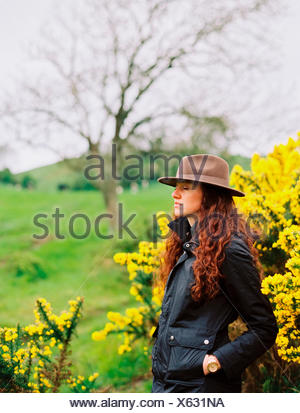 A woman with long curly hair wearing a hat, by a flowering gorse bush.