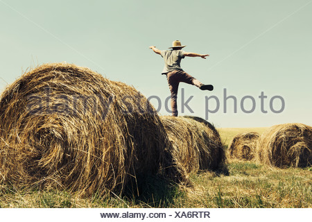 Back view of a man balancing on one leg on top of a hay bale.