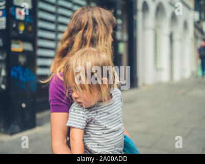 Eine junge tausendjährigen Mutter sitzt auf dem Bürgersteig mit einem traurigen Kind in ihrem Schoß Stockfoto