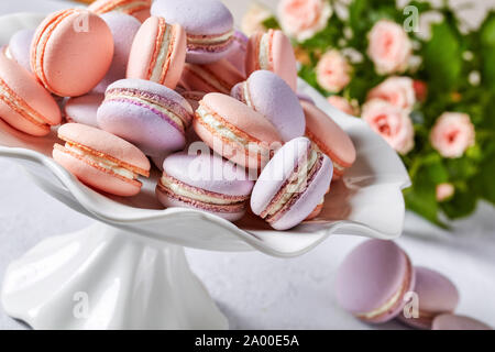 Pink und Lavendel macarons auf einem Porzellan Kuchen stand mit schönen Blumenstrauß aus Rosen im Hintergrund, horizontale Ansicht, Nahaufnahme, Makro Stockfoto