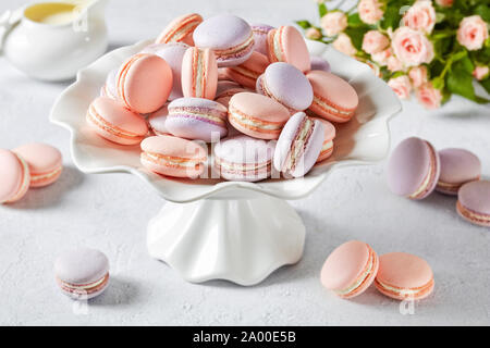 Pink und Lavendel französische Macarons auf einem Porzellan Kuchen stand mit schönen Blumenstrauß aus Rosen und Kaffeesahne im Hintergrund, horizontale Ansicht, close-up Stockfoto