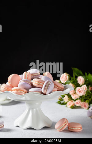 Pink und Lavendel macarons auf einem weißen Kuchen stand mit schönen Blumenstrauß aus Rosen auf dem schwarzen Hintergrund, vertikale Ansicht, close-up Stockfoto