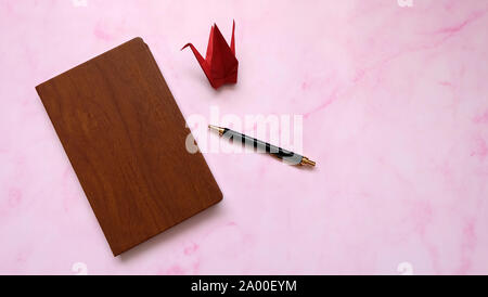 Ein Notebook mit Holzverkleidung, mit einem Kugelschreiber und einem roten Papier Kran neben ihm. Rosa Marmor Hintergrund. Stockfoto