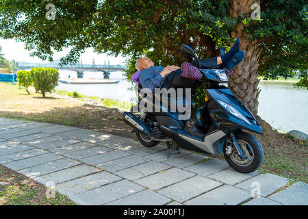 Kaohsiung, Taiwan: Alter Mann schlafen auf seinem Roller in einem Park im Schatten einer Palme, mit einem See im Hintergrund Stockfoto