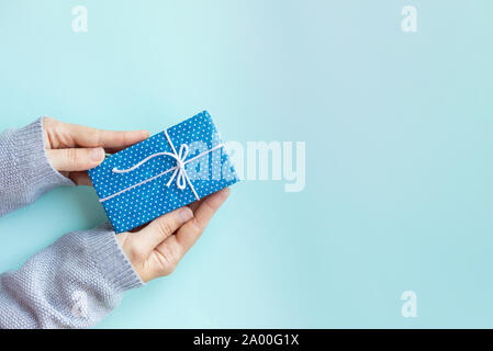Frau Hände in gemütlichen grau Pullover holding Geschenkbox. Kleines Geschenk in polkadot Blau craft Papier mit Bogen auf blauem Hintergrund eingerichtet. Kopieren Sie Platz. Stockfoto